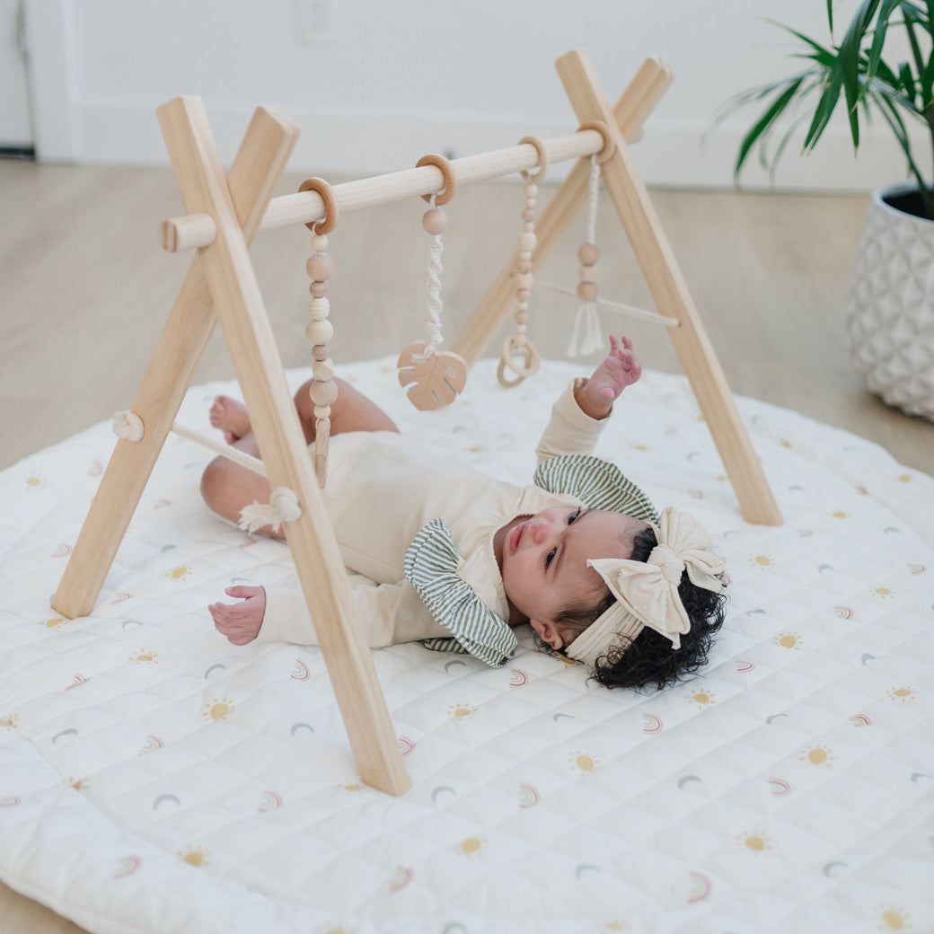 Baby laying on a mat underneath the Poppyseed Play Wood Baby Gym + Toys in -- Color_Natural _ Macrame