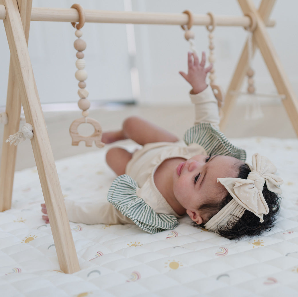 Baby with a bow playing with Poppyseed Play Wood Baby Gym + Toys in -- Color_Natural _ Macrame
