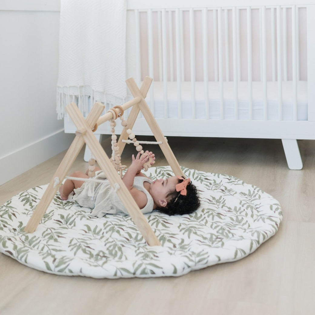 Baby laying on a mat in front of a white crib playing with Poppyseed Play Wood Baby Gym + Toys in -- Color_Natural _ Macrame
