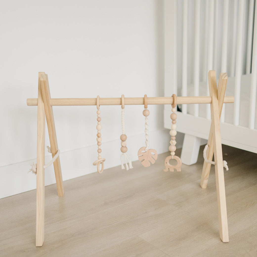 Poppyseed Play Wood Baby Gym + Toys in front of a white crib in -- Color_Natural _ Macrame