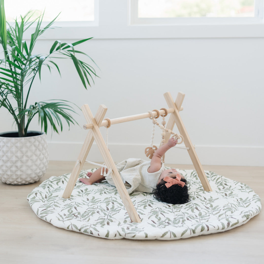 Baby laying on a leaf mat playing with the Poppyseed Play Wood Baby Gym + Toys in -- Color_Natural _ Macrame
