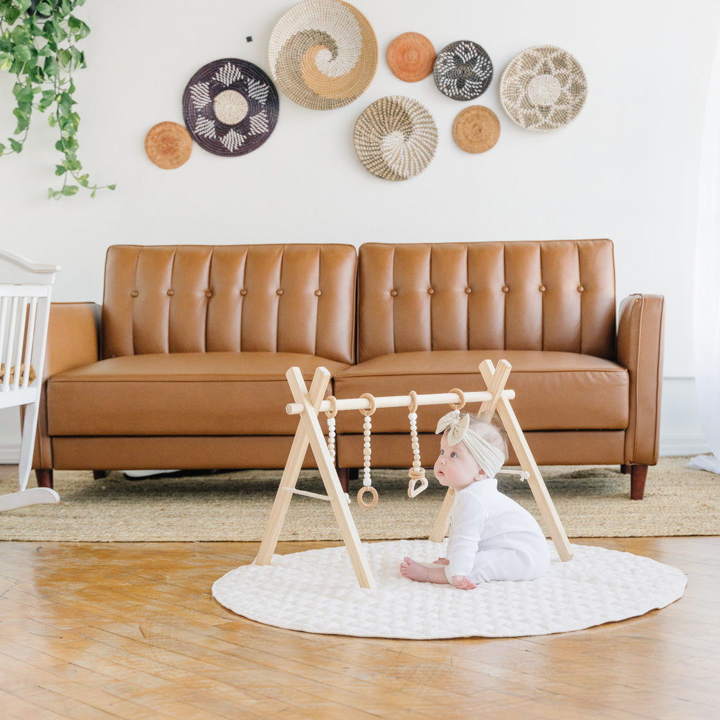 Baby in front of a couch with the Poppyseed Play Wood Baby Gym + Toys in -- Color_Natural _ White