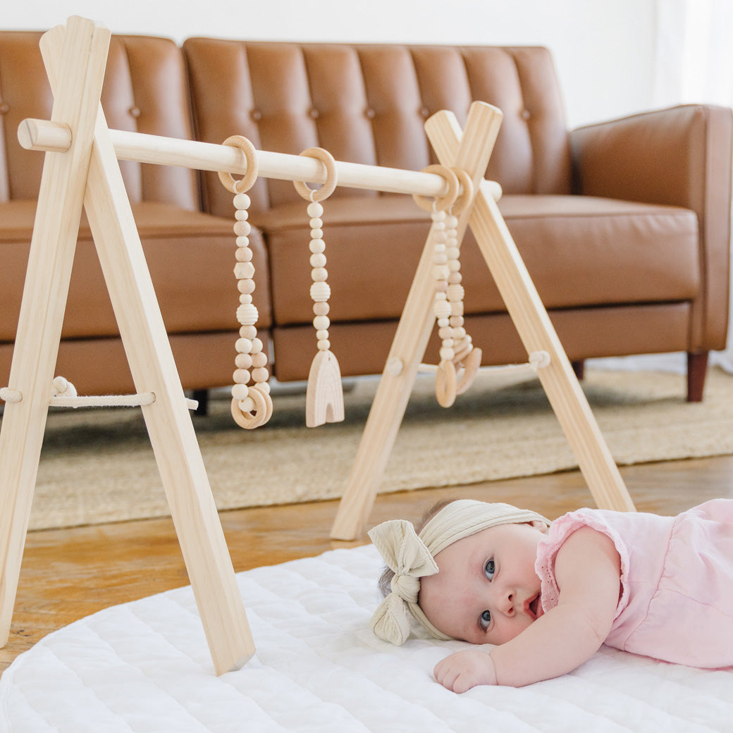 Baby laying on a mat underneath the Poppyseed Play Wood Baby Gym + Toys in -- Color_Natural _ Wood
