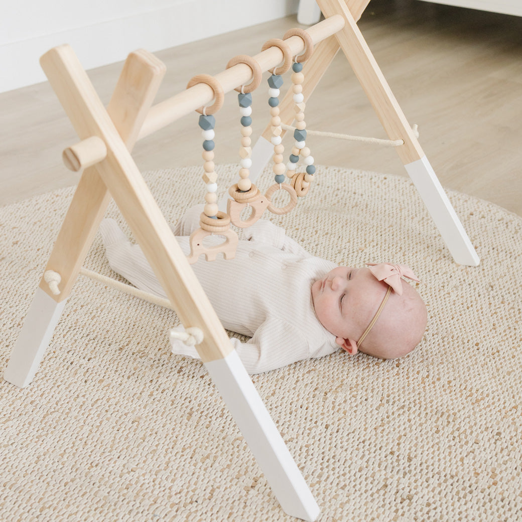 Baby with a bow underneath the Poppyseed Play Wood Baby Gym + Toys in -- Color_White _ Gray