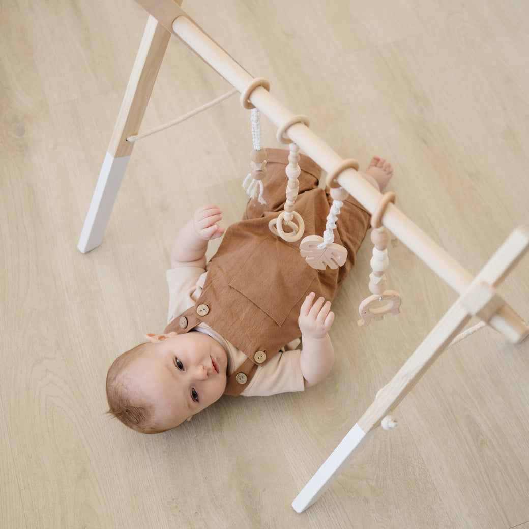 Baby in overalls playing with Poppyseed Play Wood Baby Gym + Toys in -- Color_White _ Macrame