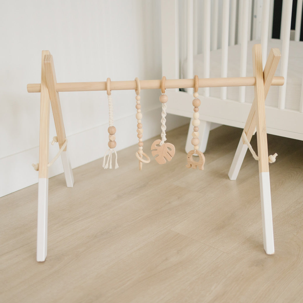 Poppyseed Play Wood Baby Gym + Toys in front of a white crib in -- Color_White _ Macrame