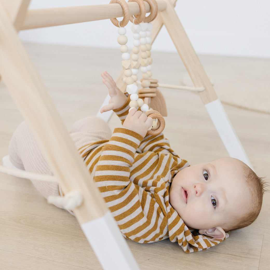 Baby holding the toys of Poppyseed Play Wood Baby Gym + Toys in -- Color_White _ White