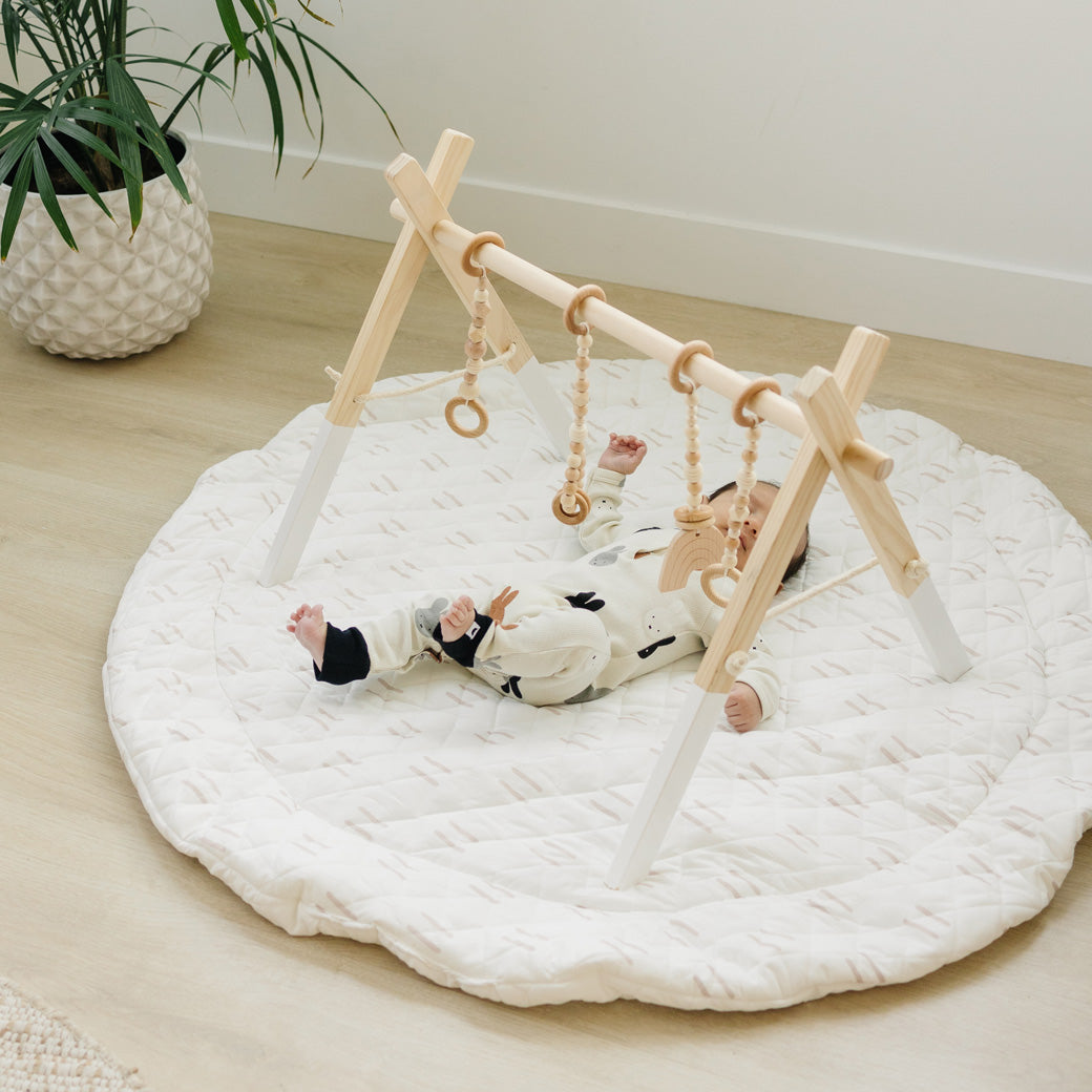 Baby playing on a mat with the Poppyseed Play Wood Baby Gym + Toys in -- Color_White _ Wood