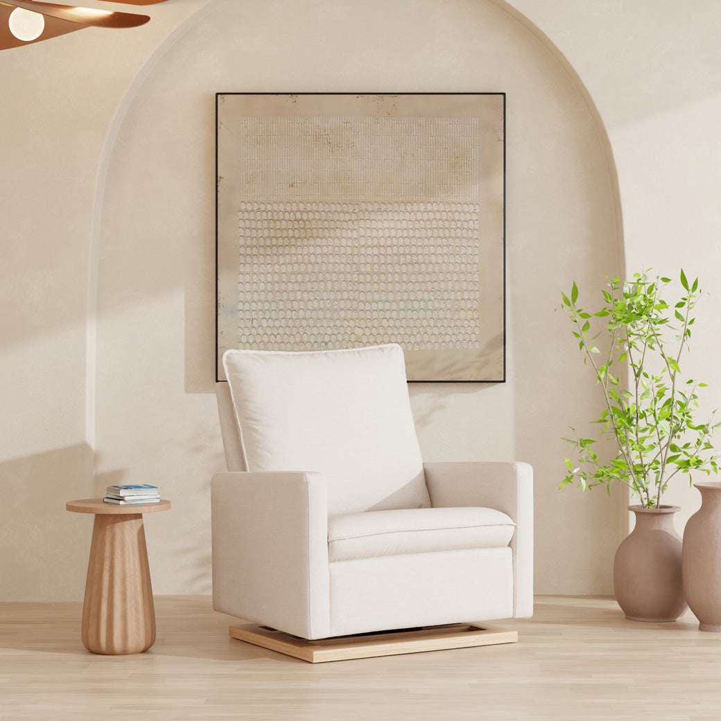 Babyletto Cali Pillowback Chair-and-a-Half Glider in a light room next to a coffee table and a plant  in -- Color_Performance Cream Eco-Weave with Light Wood Base