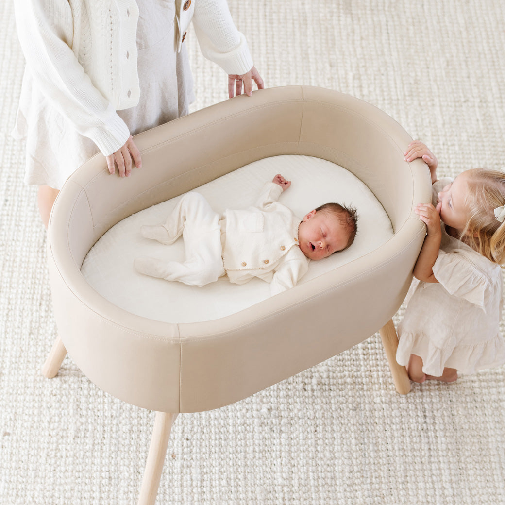 Mom and toddler looking at baby laying in Babyletto GATHRE Capsule Bassinet in -- Color_Millet