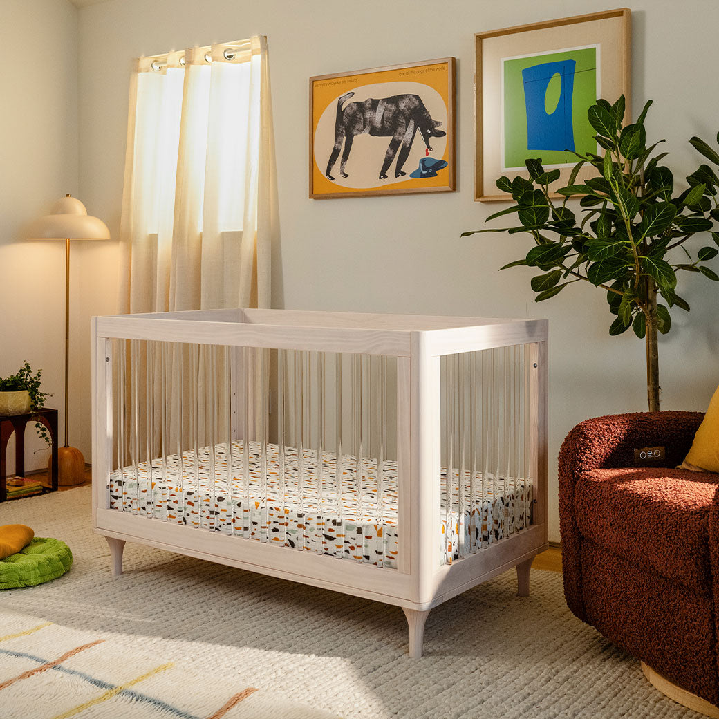 The Babyletto Lolly 3-in-1 Convertible Crib next to a window, recliner, and plant  in -- Color_Washed Natural / Acrylic