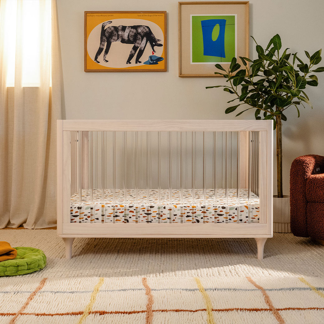 Front view of The Babyletto Lolly 3-in-1 Convertible Crib next to a window and recliner in -- Color_Washed Natural / Acrylic