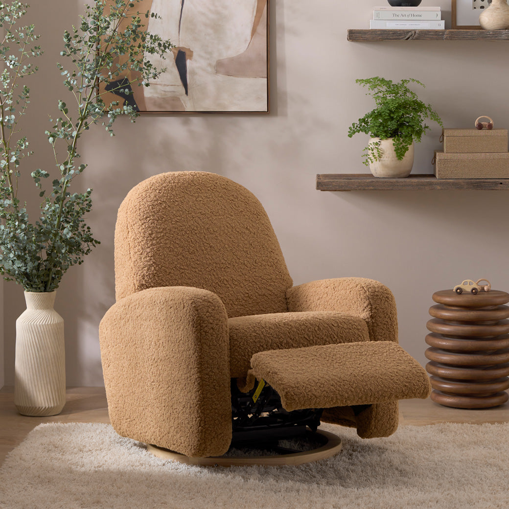 The reclined Babyletto Nami Glider Recliner next to a plant and shelf  in -- Color_Cortado Shearling with Light Wood Base
