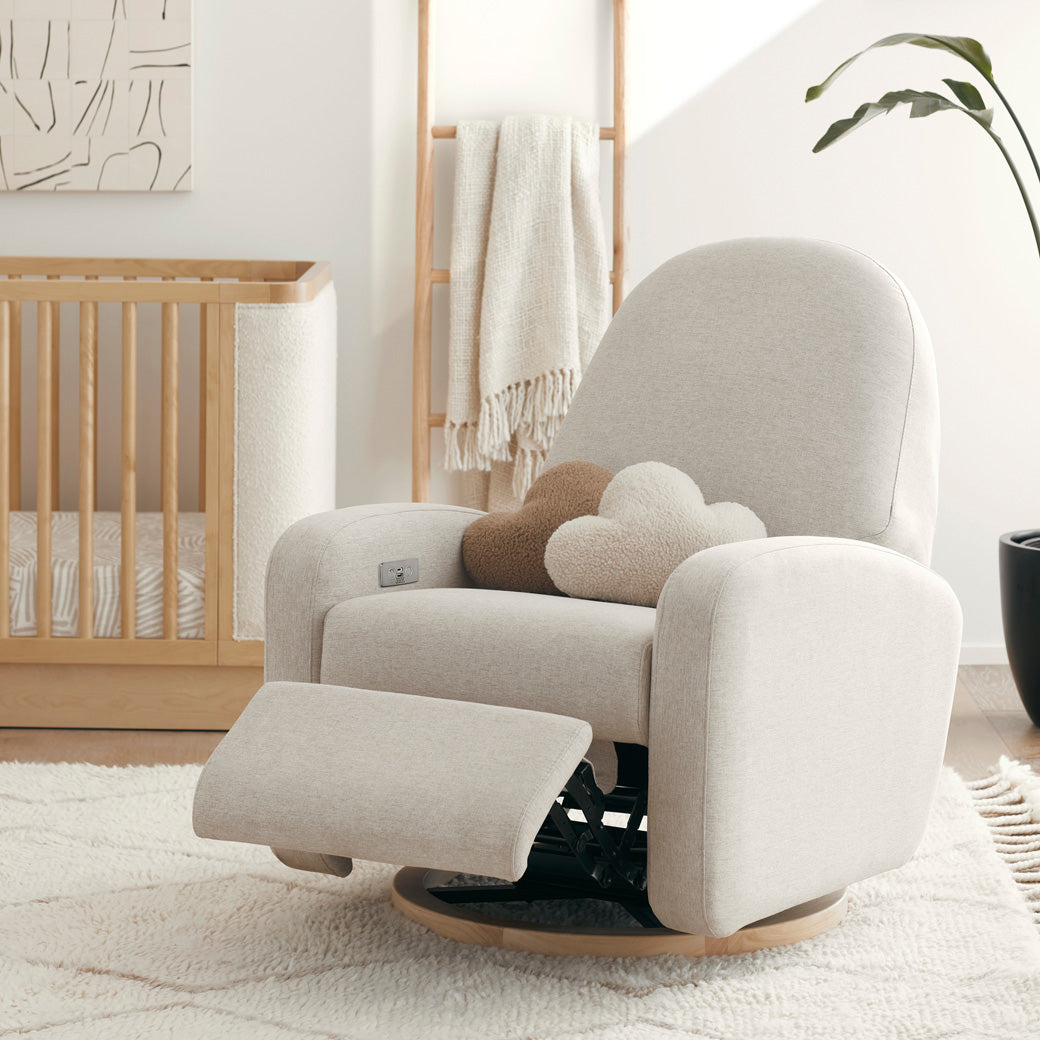 The Babyletto Nami Glider Recliner with footrest up next to a crib in -- Color_Performance Beach Eco-Weave with Light Wood Base