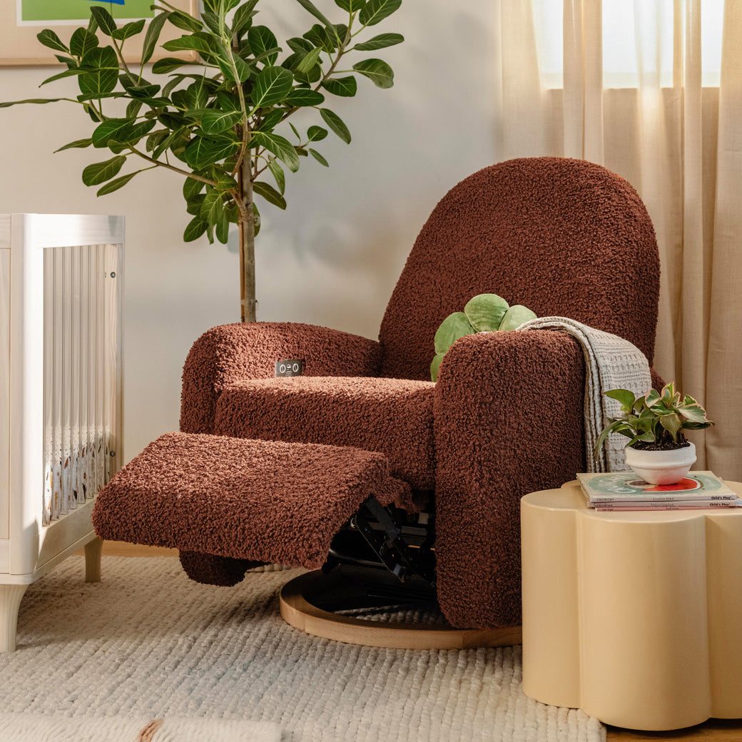 The Babyletto Nami Glider Recliner with footrest up next to a crib and coffee table  in -- Color_Rouge Teddy Loop with Light Wood Base
