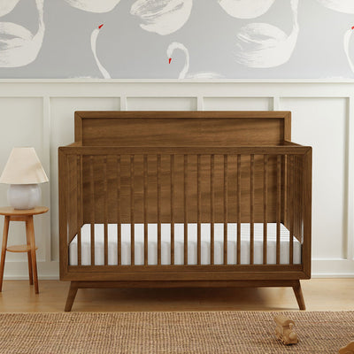 Babyletto's Palma 4-in-1 Convertible Crib next to a lamp in -- Color_Natural Walnut