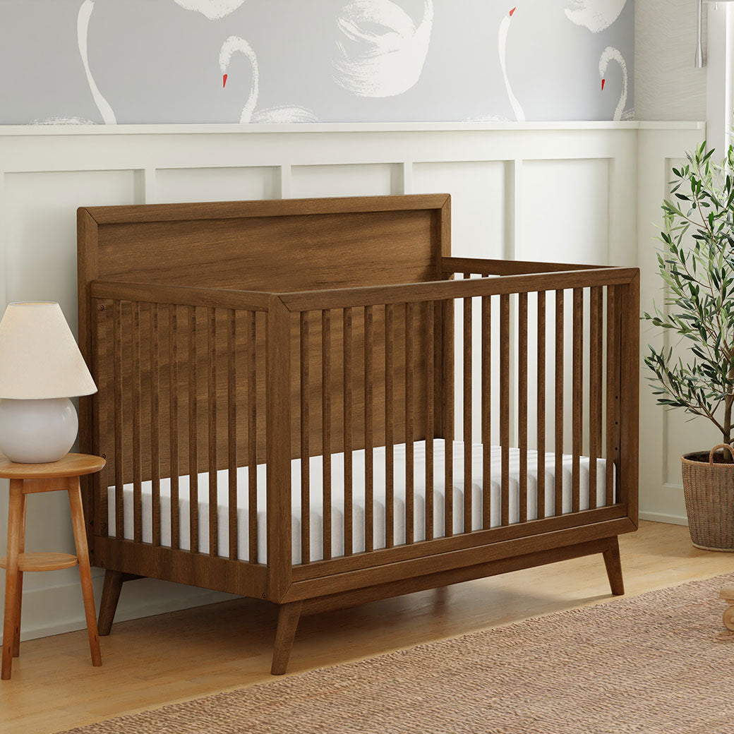 Side view of Babyletto's Palma 4-in-1 Convertible Crib next to a lamp in -- Color_Natural Walnut
