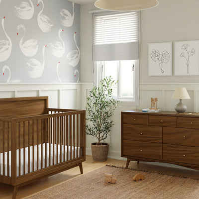 Babyletto's Palma 7-Drawer Assembled Double Dresser next to a crib and plant  in -- Color_Natural Walnut