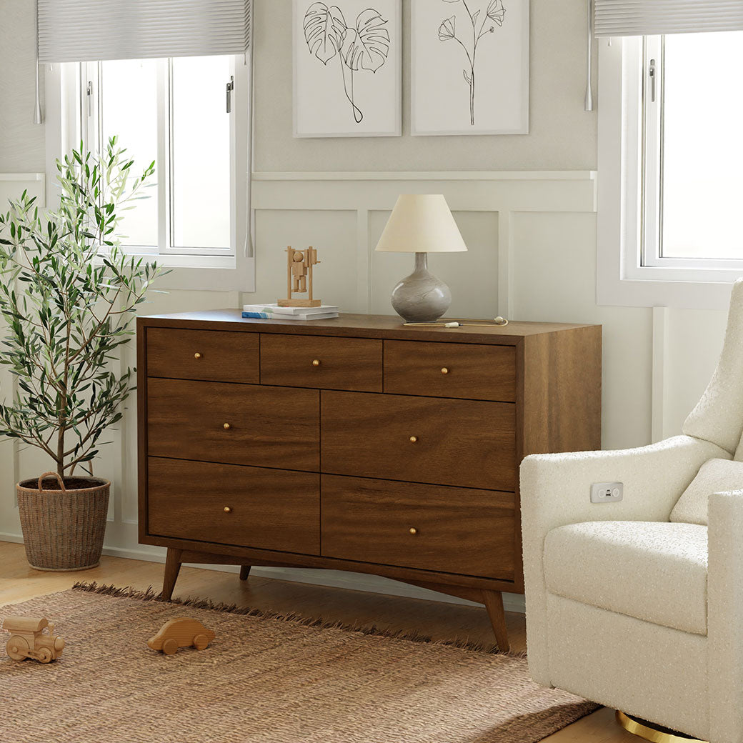 Babyletto's Palma 7-Drawer Assembled Double Dresser next to a recliner and plant  in -- Color_Natural Walnut
