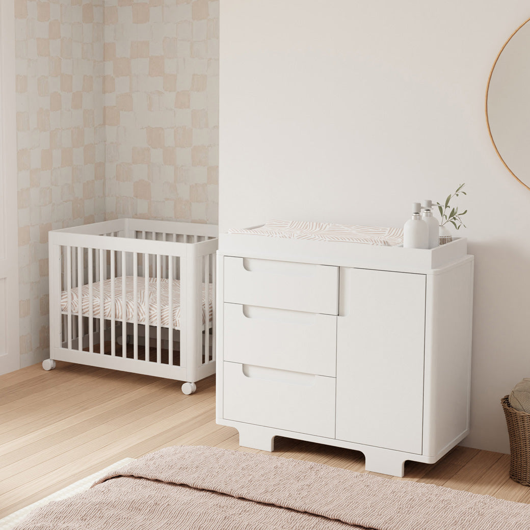 Babyletto Yuzu 3-Drawer Changer Dresser next to a crib and basket  in -- Color_White