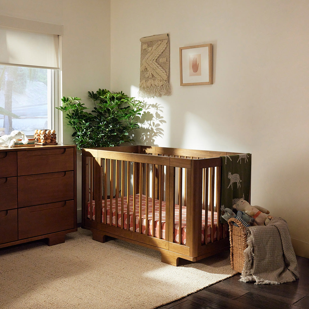 Babyletto's Yuzu 8-In-1 Convertible Crib next to a dresser and basket  in -- Color_Natural Walnut