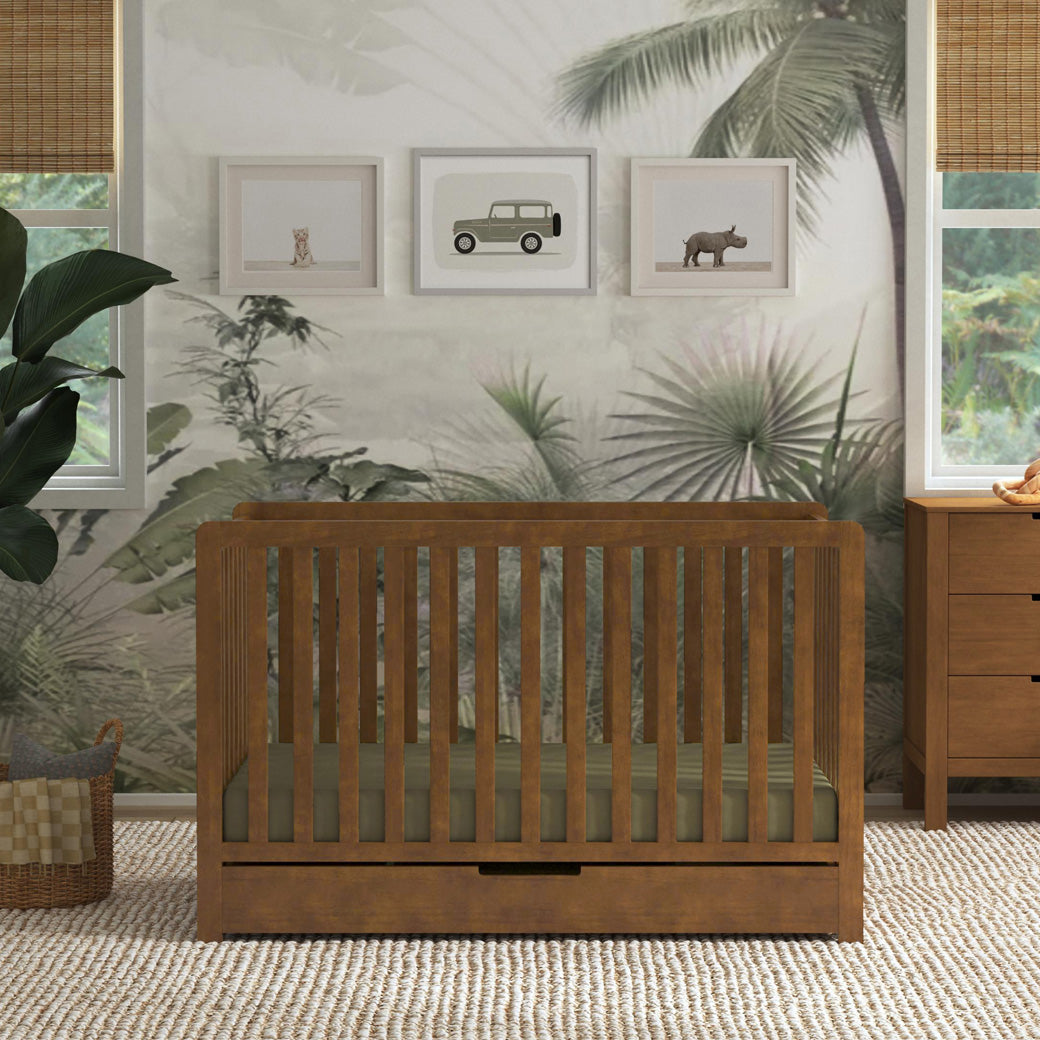 Front view of Carter's by DaVinci Colby 4-in-1 Convertible Crib with Trundle Drawer next to a dresser and plant in -- Color_Walnut