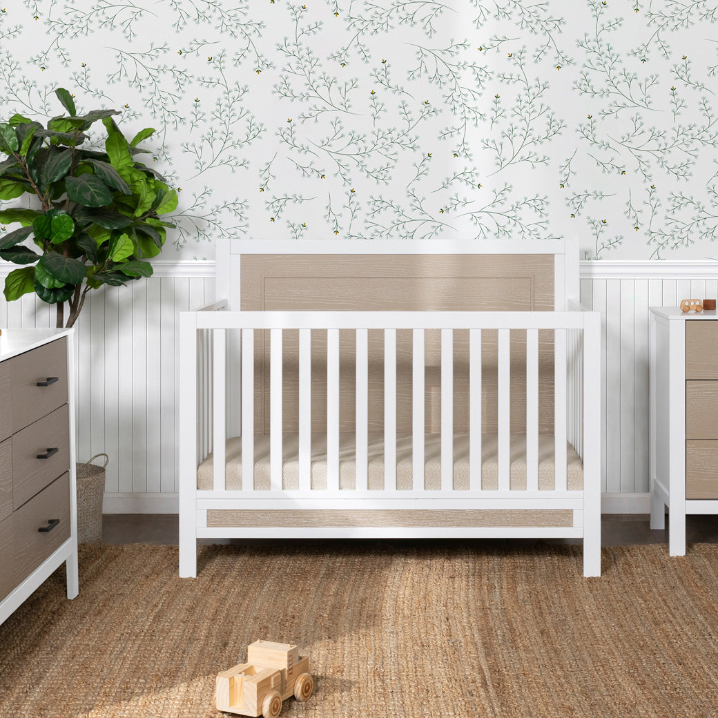 Front view of Carter's by DaVinci Radley 4-in-1 Convertible Crib next to two dressers and a plant  in -- Color_White/Coastwood