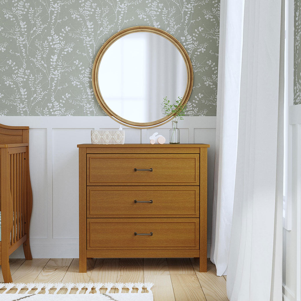 The DaVinci Charlie 3-Drawer Dresser under a mirror next to a crib and window in -- Color_Chestnut
