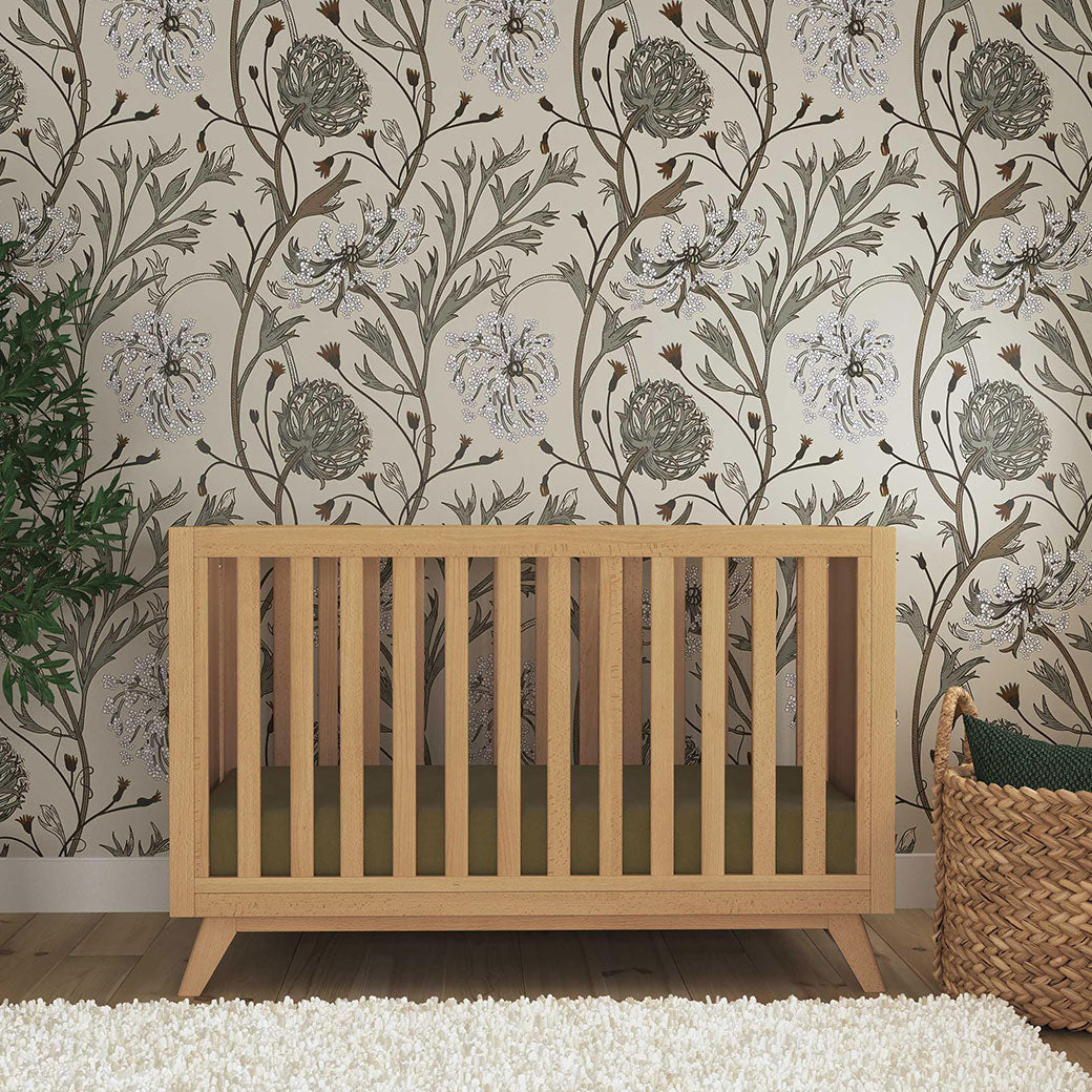 Front view of the DaVinci Otto 3-in-1 Convertible Crib next to a plant and basket in -- Color_Honey _ Wood