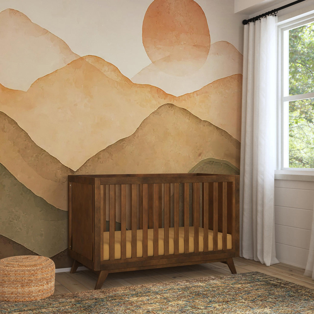 Angled view of DaVinci Otto 3-in-1 Convertible Crib in a sunset and mountains room in -- Color_Walnut _ Wood