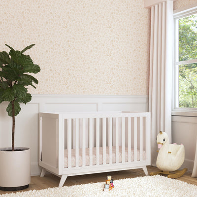 Angled view of DaVinci Otto 3-in-1 Convertible Crib next to a plant and window in -- Color_White _ Wood