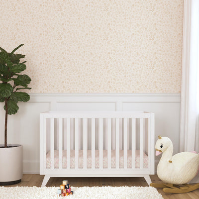 Front view of DaVinci Otto 3-in-1 Convertible Crib next to a plant and a swan rocker   in -- Color_White _ Wood