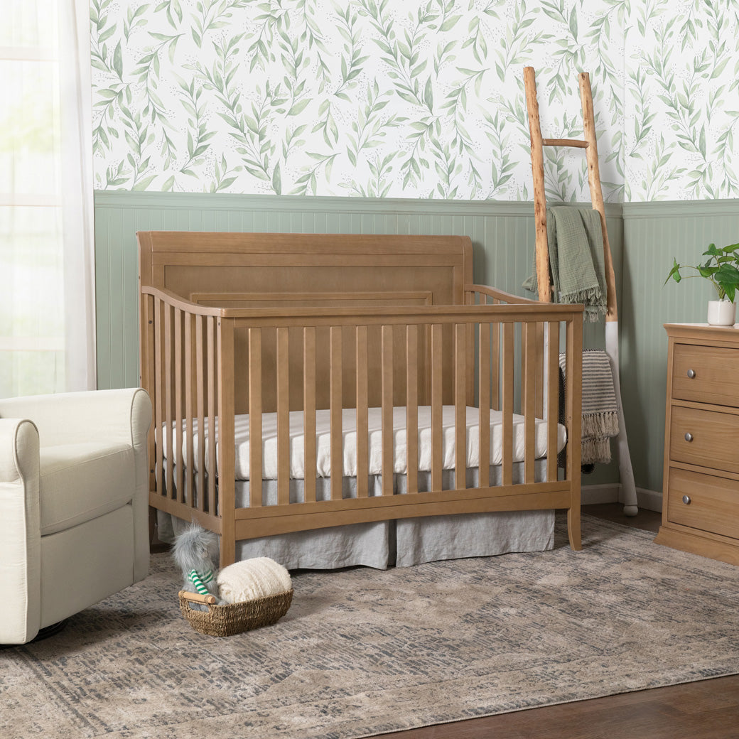 DaVinci Anders 4-in-1 Convertible Crib next to a recliner and dresser in -- Color_Hazelnut