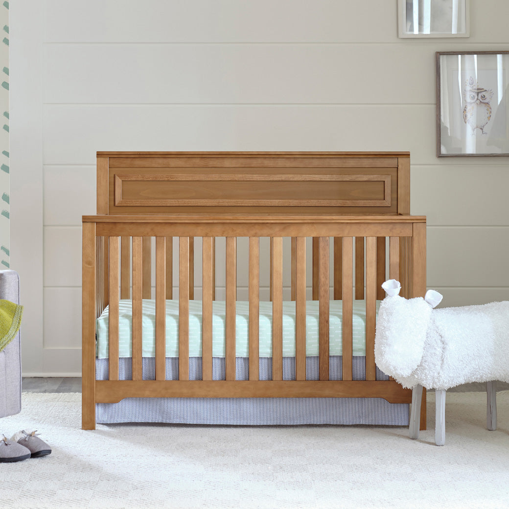Front view of DaVinci Autumn 4-in-1 Convertible Crib next to a sheep stool  in -- Color_Chestnut