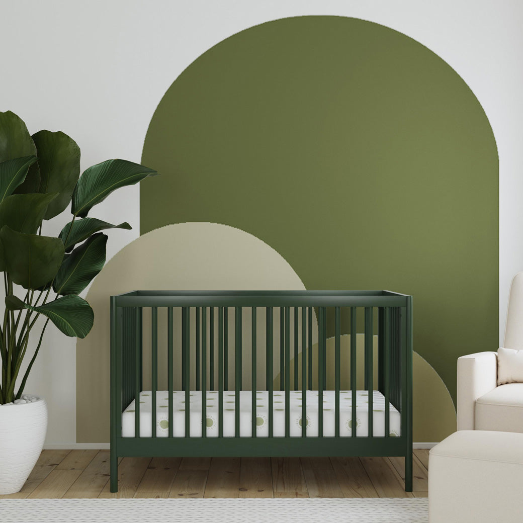Front view of DaVinci Birdie 3-in-1 Convertible Crib next to a plant and recliner in -- Color_Forest Green