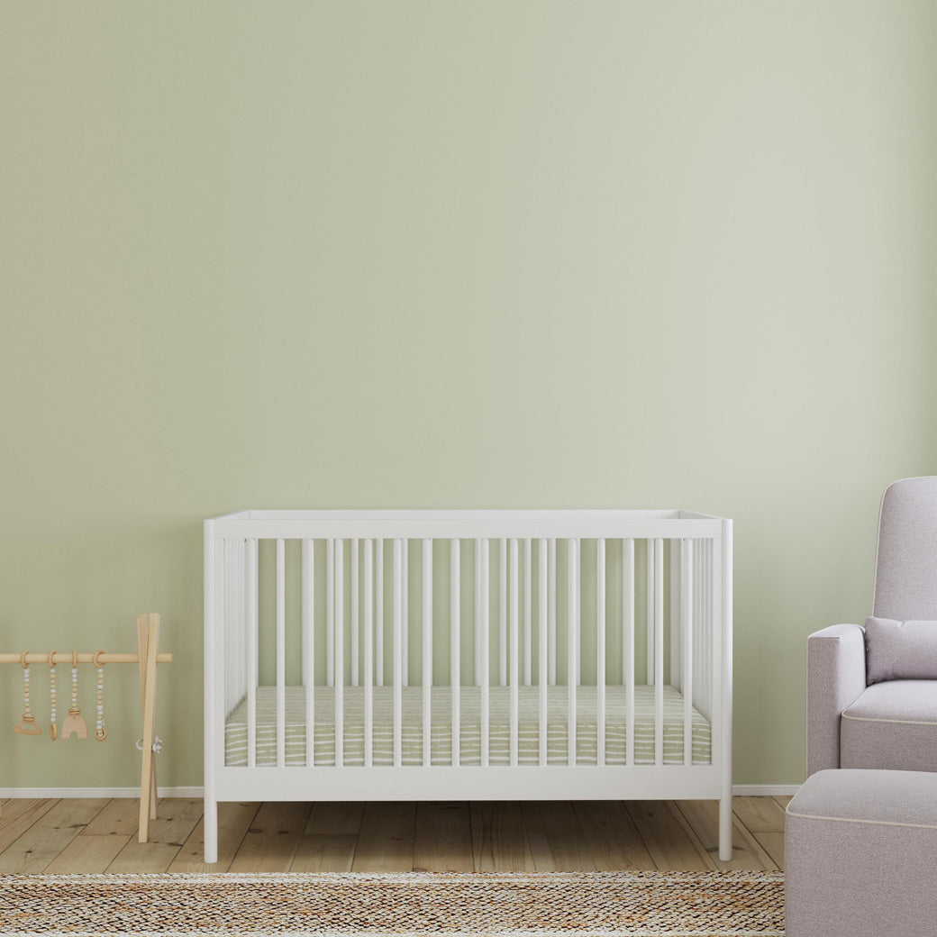 Front view of DaVinci Birdie 3-in-1 Convertible Crib next to a play toy bar and recliner  in -- Color_White