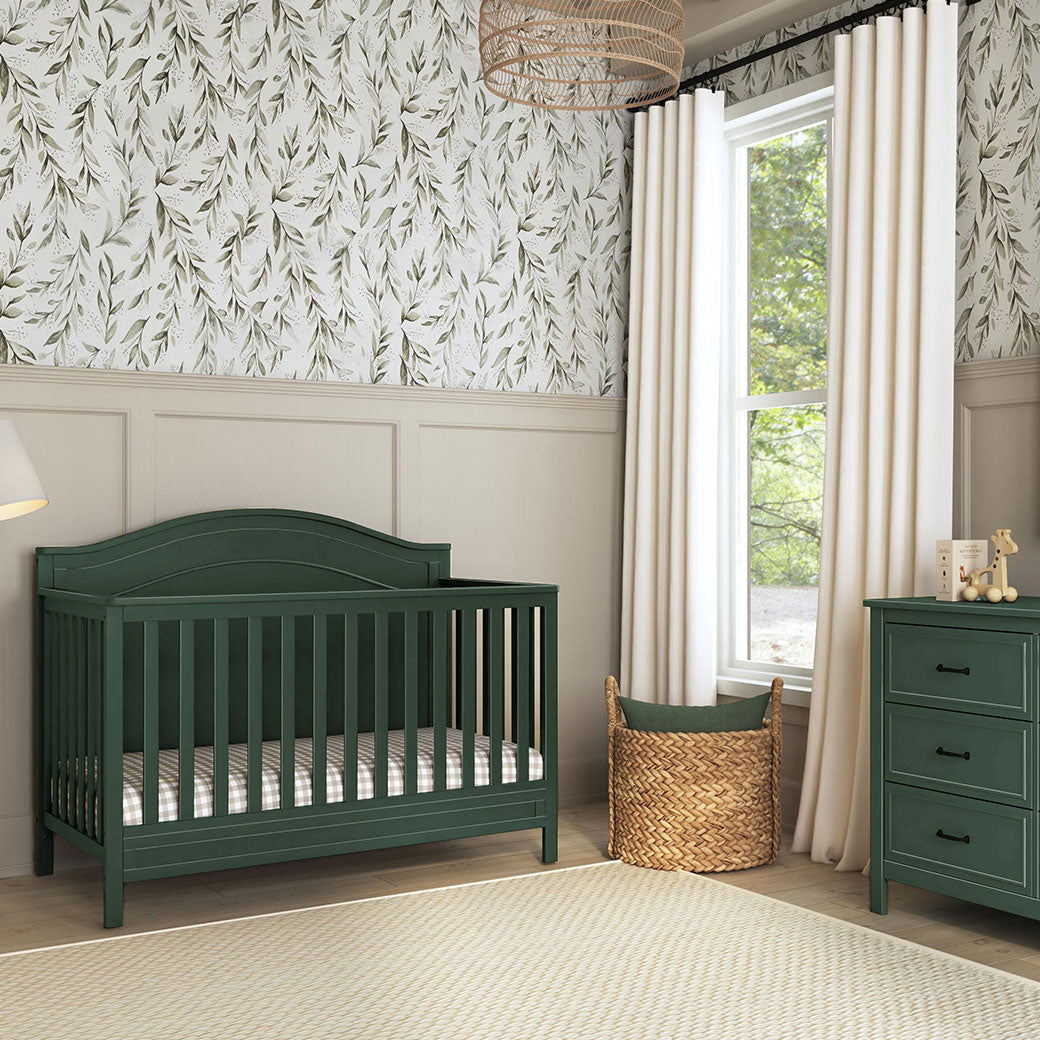 Front view of The DaVinci Charlie 4-in-1 Convertible Crib next to a basket, window, and dresser in -- Color_Forest Green