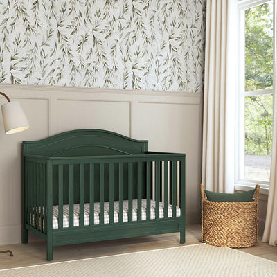 Front view of The DaVinci Charlie 4-in-1 Convertible Crib next to a basket and window in -- Color_Forest Green
