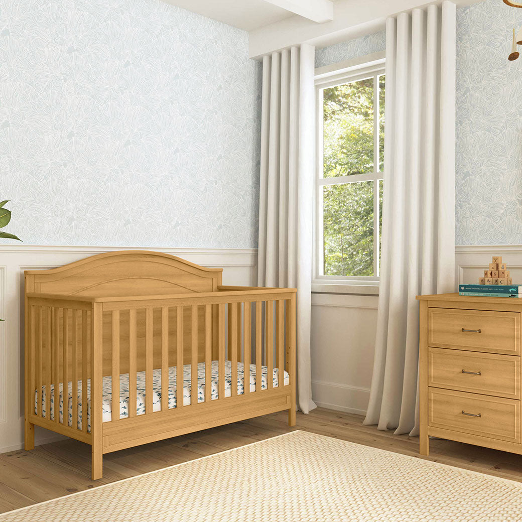 The DaVinci Charlie 4-in-1 Convertible Crib next to a window and dresser in -- Color_Honey