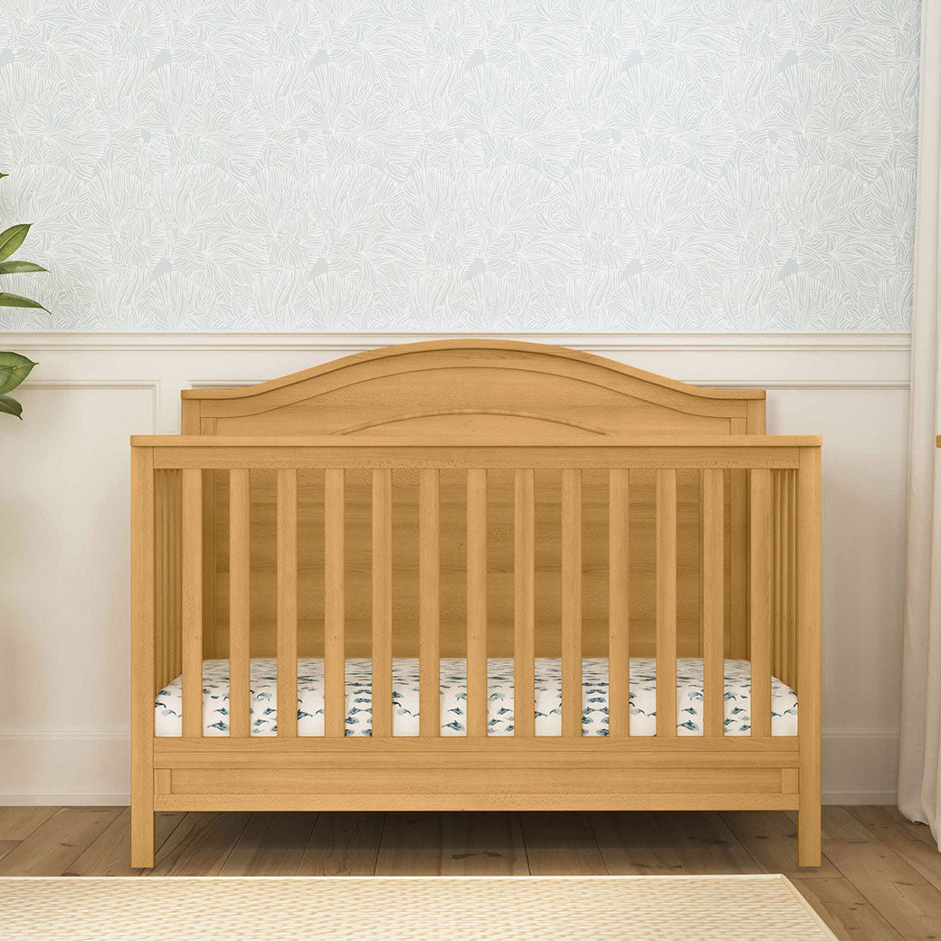 Front view of The DaVinci Charlie 4-in-1 Convertible Crib next to a plant in -- Color_Honey
