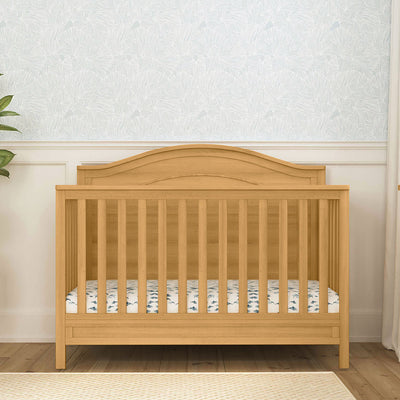 Front view of The DaVinci Charlie 4-in-1 Convertible Crib next to a plant in -- Color_Honey