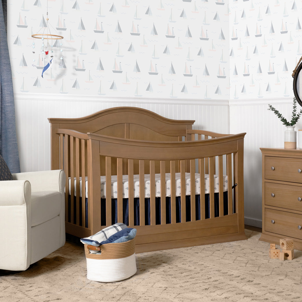 DaVinci Meadow 4-in-1 Convertible Crib next to a recliner and dresser  in -- Color_Hazelnut