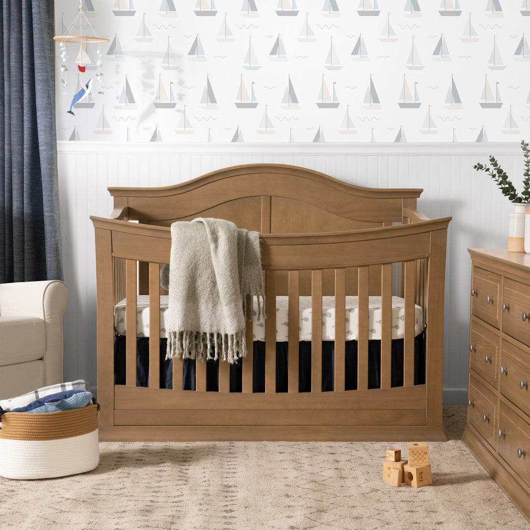 Front view of DaVinci Meadow 4-in-1 Convertible Crib next to a recliner and dresser in -- Color_Hazelnut