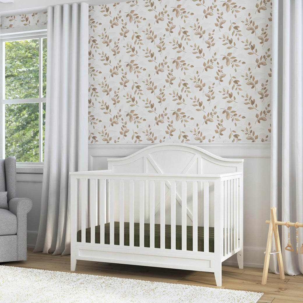 DaVinci Sawyer Farmhouse 4-in-1 Convertible Crib next to a window and recliner  in -- Color_Heirloom White