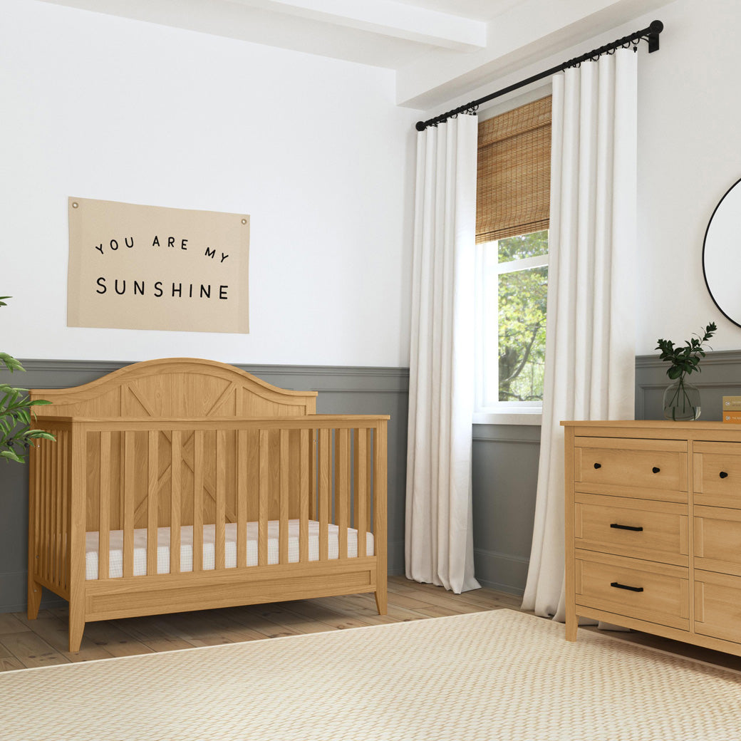 DaVinci Sawyer Farmhouse 4-in-1 Convertible Crib next to a window and dresser  in -- Color_Honey