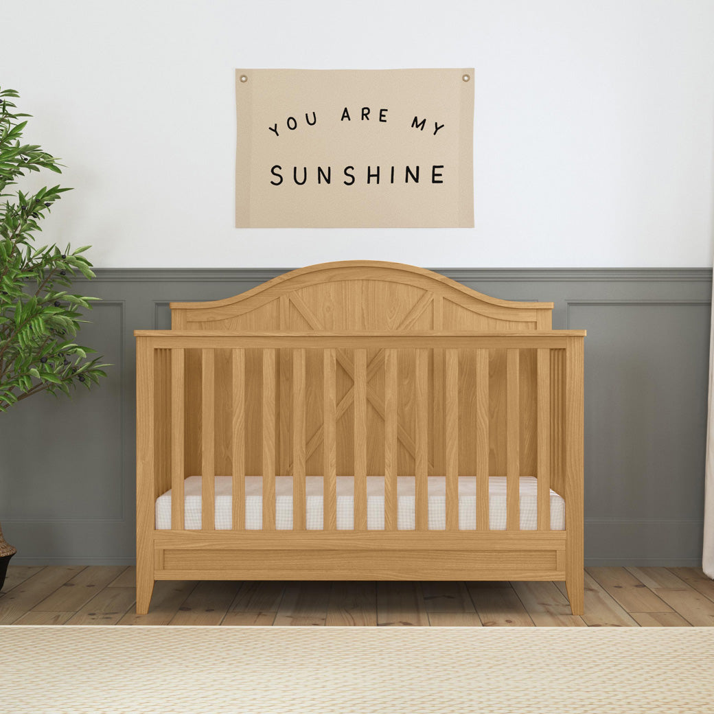 Front view of DaVinci Sawyer Farmhouse 4-in-1 Convertible Crib next to a plantin -- Color_Honey