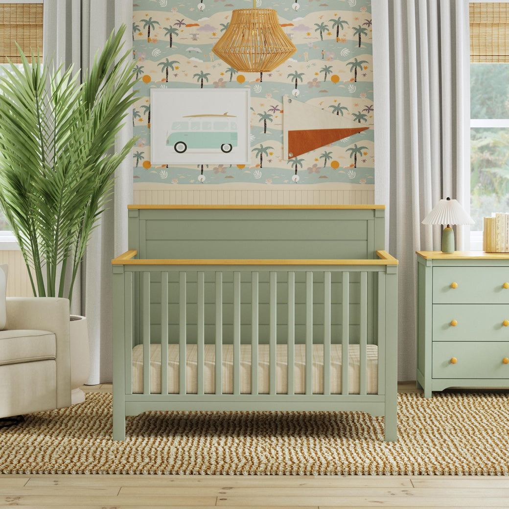 Front view of DaVinci Shea 4-in-1 Convertible Crib next to recliner and dresser in -- Color_Light Sage/Honey