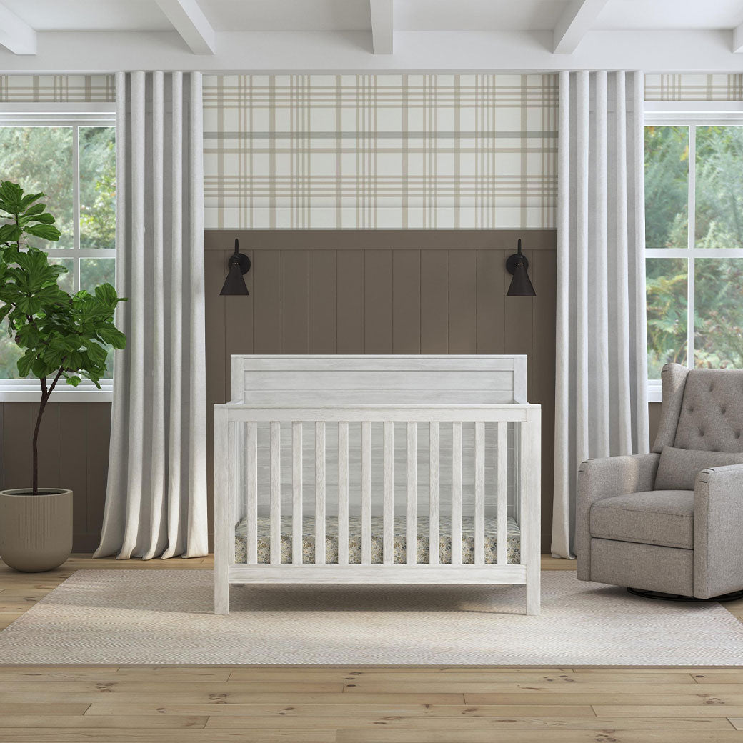 Front view of DaVinci Fairway 4-in-1 Convertible Crib next to a recliner and window in -- Color_Cottage White