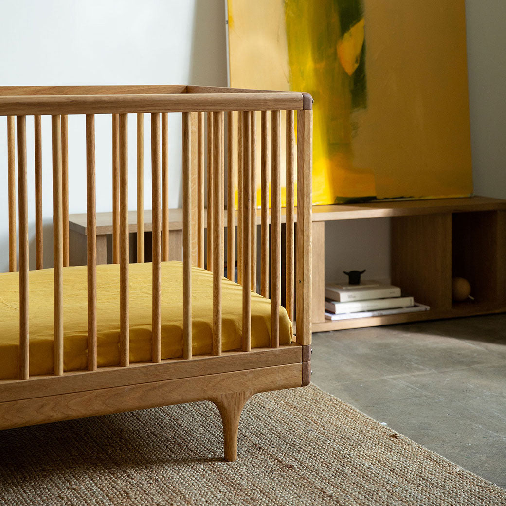 Kalon Caravan Crib in a room next to a yellow painting  in -- Color_White Oak