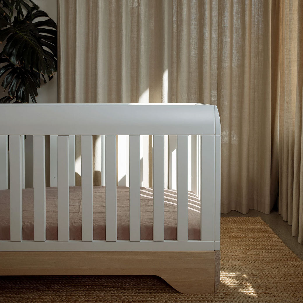 Right side view of the Kalon Echo Crib in front of a curtain and a plant  in -- Color_Salt
